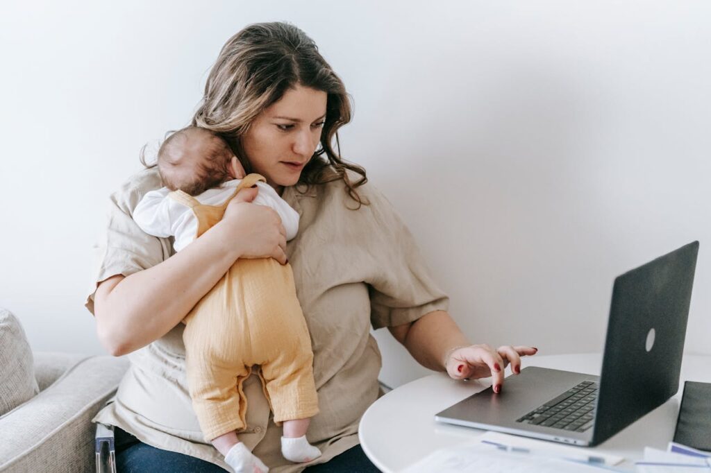 pexels-photo-7282818 Concentrated young female freelancer embracing newborn while sitting at table and working remotely on laptop at home